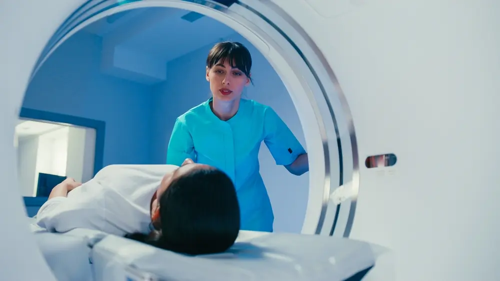 A female medic in a teal uniform supports a patient during an MRI scan, speaking gently and maintaining eye contact through the scanner opening.